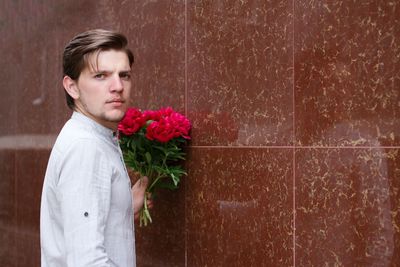 Side view of handsome young man with flowers standing by wall