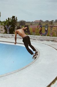 Full length of shirtless man skateboarding in swimming pool against sky