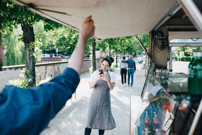 Female owner photographing through smart phone while male coworker opening shade of food truck