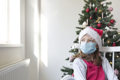 Cute girl wearing mask looking away while sitting against christmas tree at home