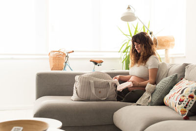 Pregnant mid adult woman with bag sitting on sofa against window at home