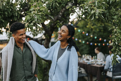 Cheerful young woman with hand on shoulder of male friend standing in back yard at dinner party