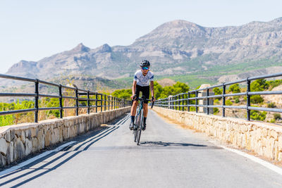 Full length of active female bicyclist in helmet and sportswear riding bike on asphalt road near mountain ridge in summer day