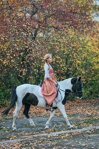 Side view of young woman riding horse on pathway against trees during autumn