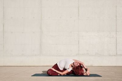 Barefooted woman with with closed eyes in sportswear doing yoga revolved head to knee pose on mat training alone on street against concrete wall