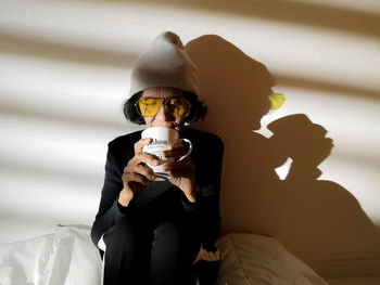 Senior woman drinking coffee while sitting against wall at home