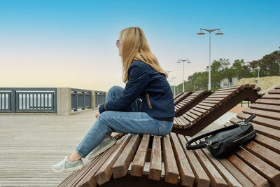 Blonde woman on empty city embankment rests sitting on sun lounger and looks into the distance.