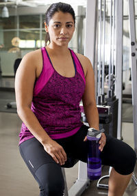 Portrait of woman holding bottle while sitting on exercise equipment in gym