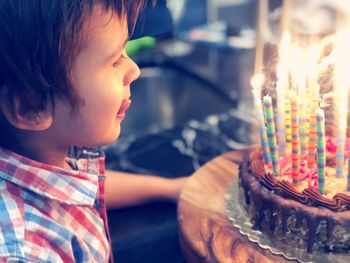 Side view of cute boy sticking out tongue while looking at illuminated birthday cake at home
