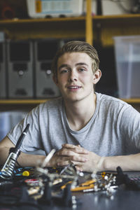 Portrait of confident male teenage student sitting with science project at desk in classroom at high school