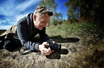 Smiling photographer photographing field through camera against sky