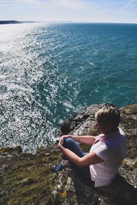 Rear view of boy sitting on retaining wall by sea against sky