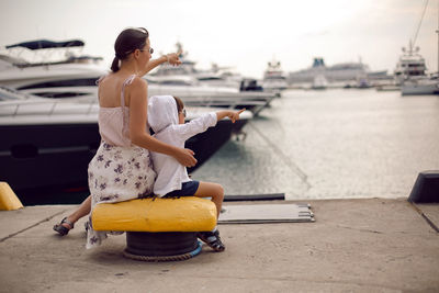 Mother in a dress with her son sitting on the pier with yachts on the sea in the summer