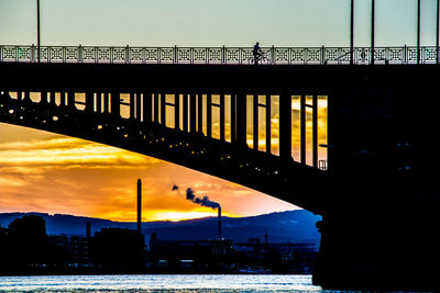 Silhouette bridge over river in city against sky during sunset