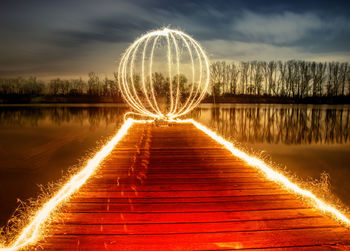 Illuminated firework display on pier over lake against sky at night