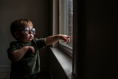 Toddler boy wearing round glasses standing by window pointing outside