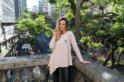 Portrait of young woman standing against trees in city