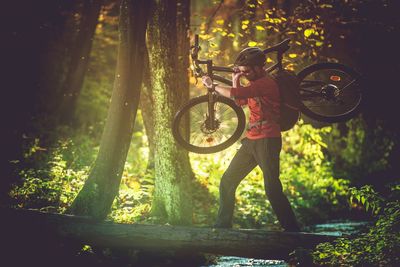 Man carrying bicycle on tree trunk over stream in forest