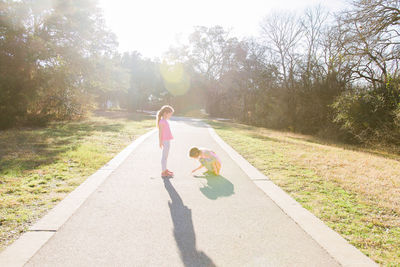 Full length of girl looking at sister drawing on footpath in park against trees