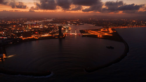 Illuminated cityscape by sea against sky at sunset