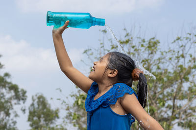 An indian girl pouring water on head from a blue plastic bottle under sky to cool down in summer