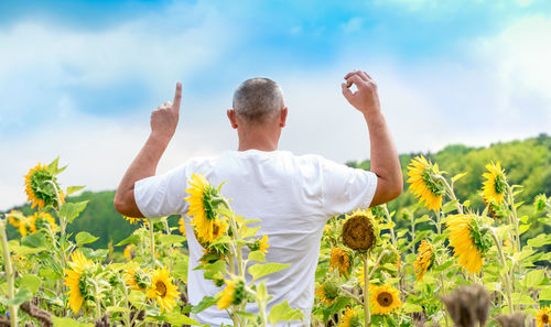 Rear view of man with arms raised gesturing while standing on sunflower field against sky