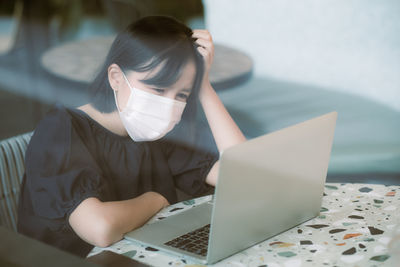 Woman with head in hands wearing while mask sitting by laptop on table