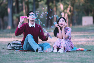 Woman enjoying while sitting besides man blowing bubbles at park