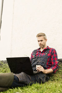 Man using laptop while sitting on grassy field against house at farm