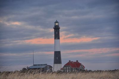 Lighthouse on field by building against sky during sunset