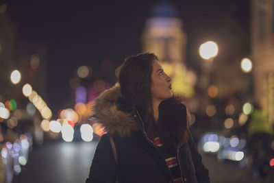 Thoughtful young woman standing on street in illuminated city at night