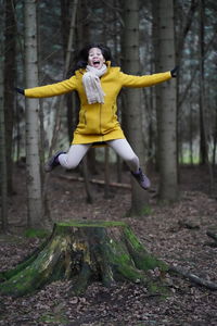 Full length of excited woman with arms outstretched jumping against trees in forest