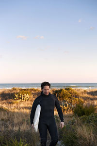 Surfer carrying surfboard while walking on grassy field against sky