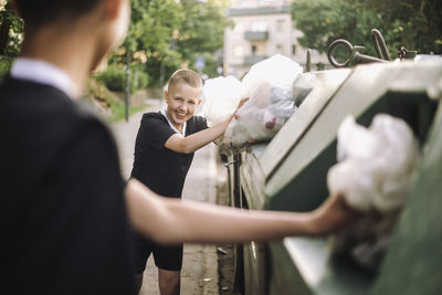 Smiling boy looking at friend while putting garbage bags in recycling bins