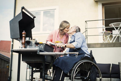 Disabled man in wheelchair kissing woman while barbecuing at yard