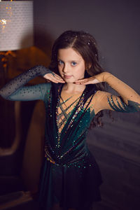 Girl in a green figure skating dress stands next to a chair and a lamp in the room