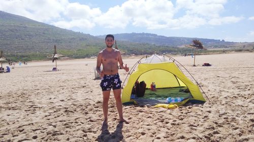 Portrait of shirtless young man camping at beach against mountains