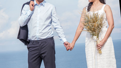 Midsection of couple holding hands while standing against sky