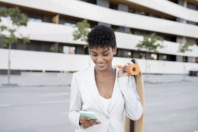 Smiling businesswoman using smart phone while carrying skateboard