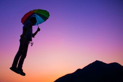 Low angle view of silhouette man with multi colored umbrella flying against clear sky during sunset