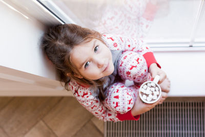 High angle portrait of cute girl holding hot chocolate sitting by windows