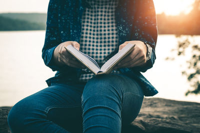 Midsection of woman holding book while sitting against lake during sunset