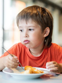 Fussy kid in red t-shirt eats pancakes with knife and fork.little boy with funny expression on face.