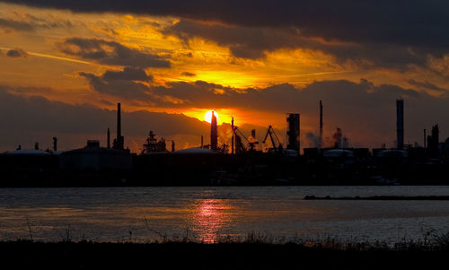 Factory at waterfront against cloudy sky during sunset
