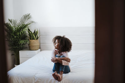 Contemplating girl looking away while sitting on bed in bedroom at home