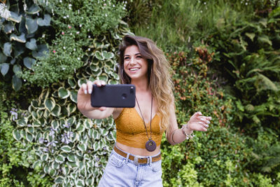 Smiling beautiful woman taking selfie from mobile phone while standing against plants at park