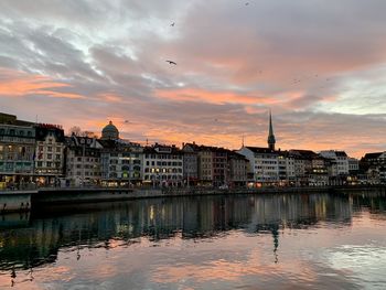 Buildings by river against sky during sunset