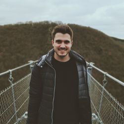 Portrait of smiling young man standing on footbridge against mountain