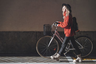 Woman wearing hat looking away while walking with bicycle against wall