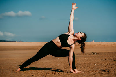 Slim female in sportswear standing on sandy beach in extended side angle pose and practicing yoga while looking up